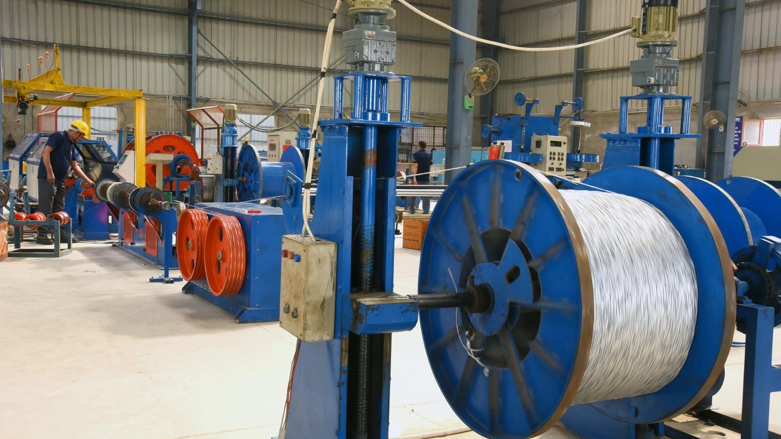 Drawn wire filling a take-up drum on the wire line at the Batra Henlay Cables plant, with an operator working at a machine behind Drawn wire filling a take-up drum on the wire line at the Batra Henlay Cables plant, with an operator working at a machine behind